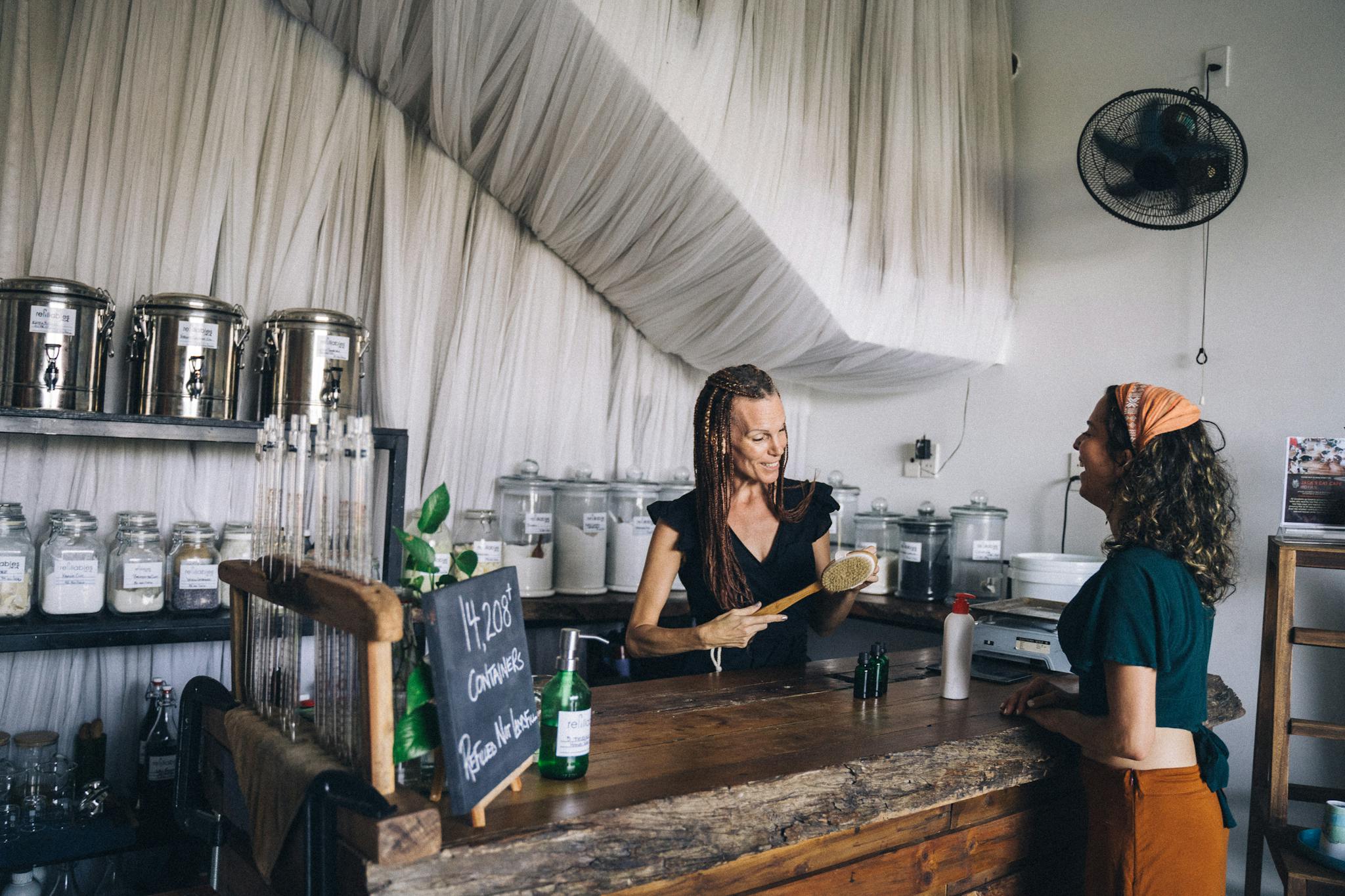 Two women engaging at an eco-friendly shop counter with refillable containers and glass jars.